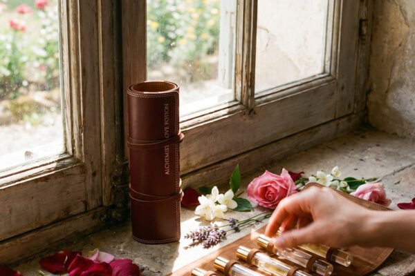 Elyamor Alchemist’s Love Potion custom perfume kit with glass vials on a leather roll near a vintage window with flowers and rose petals